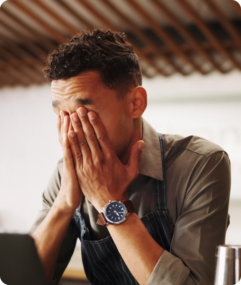 Man in an apron sitting at a table with his hands covering his face, wearing a watch